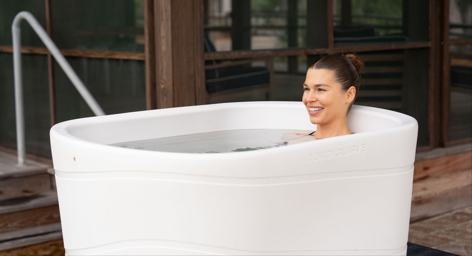 Woman sitting in a white outdoor cold tub with a wooden deck and greenery in the background